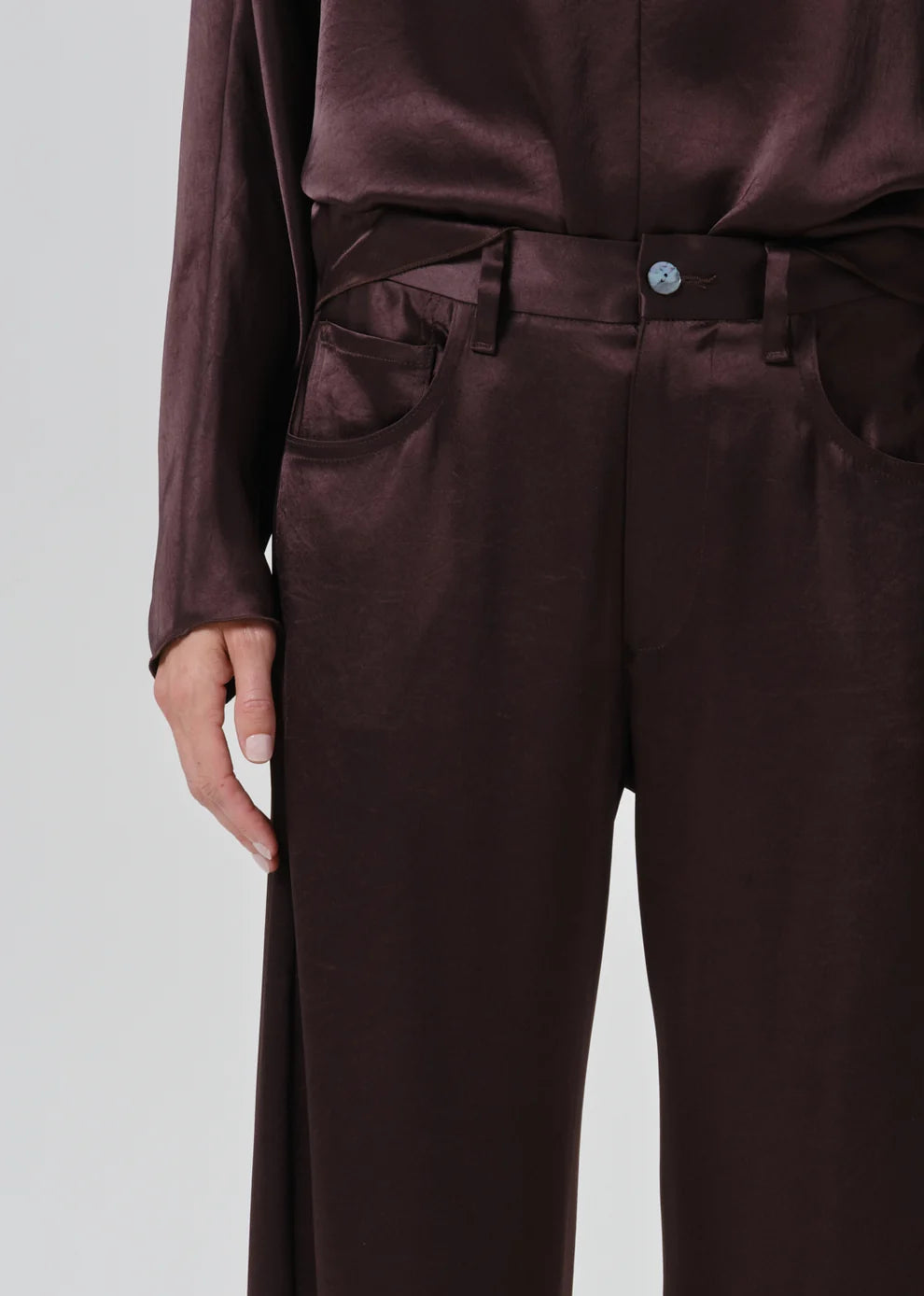 Close-up of a person wearing dark brown satin trousers with a matching top, showing the waist, belt, front pockets, and hand against a light gray background.