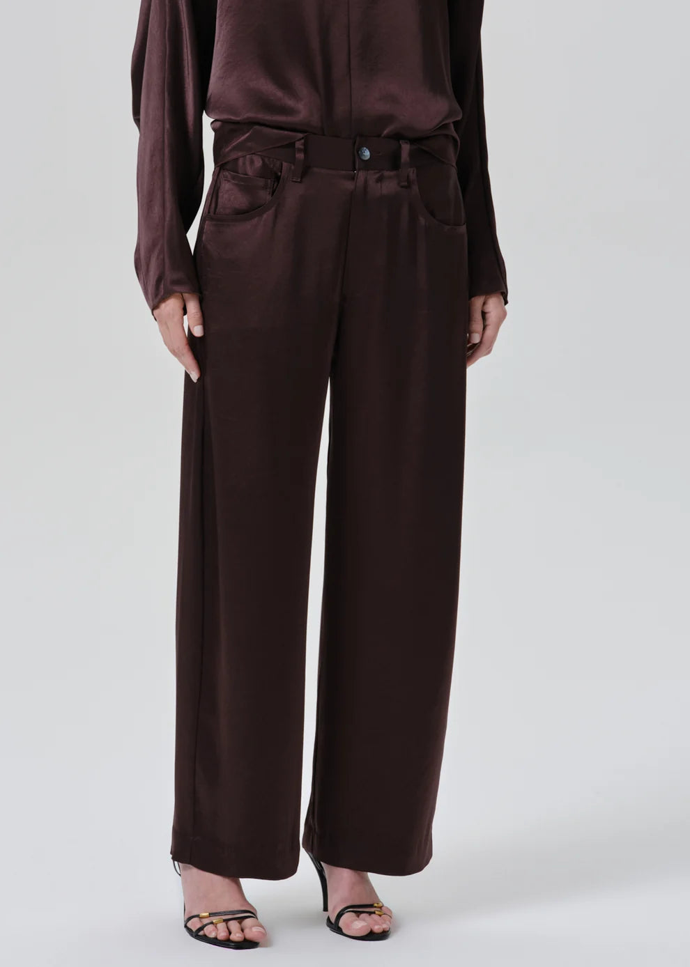 A model wearing dark brown wide-leg trousers with a matching belt and waist tie, paired with a matching dark brown satin long-sleeve top and black strappy heels, against a light gray studio background.