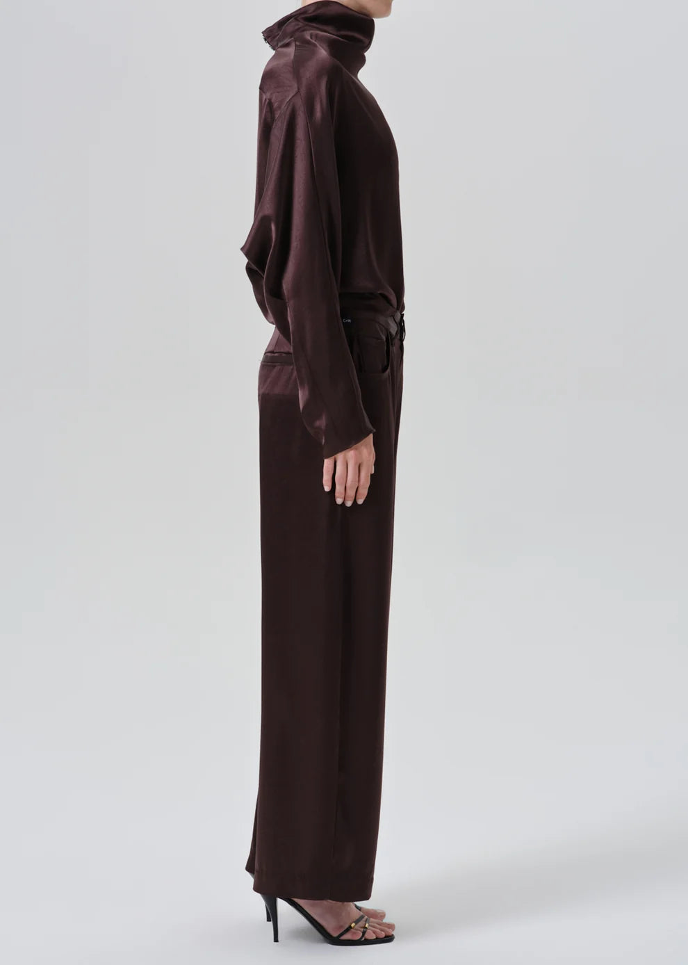 A model shown from the side wearing a dark brown satin turtleneck top and matching wide-leg trousers with a tie belt at the waist, paired with black high-heeled sandals, against a light gray studio background.