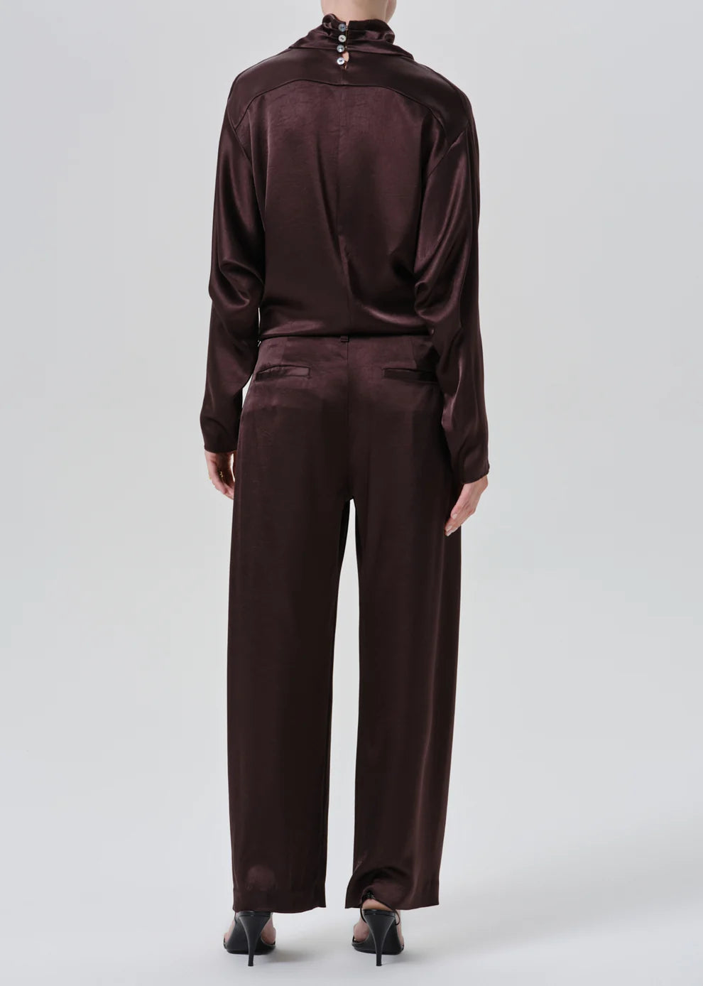 A model shown from the back wearing a matching dark plum satin tracksuit with a high-neck top and wide-leg trousers, paired with black pointed-toe heels, against a light gray studio background.
