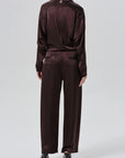 A model shown from the back wearing a matching dark plum satin tracksuit with a high-neck top and wide-leg trousers, paired with black pointed-toe heels, against a light gray studio background.