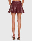 A model shown from the back wearing a short, flared faux-leather mini skirt in a deep burgundy color, paired with matching burgundy high-heeled sandals, against a light gray studio background.