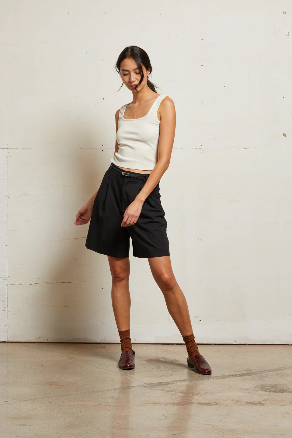 Woman wearing a white tank top and black shorts standing against a white wall front view