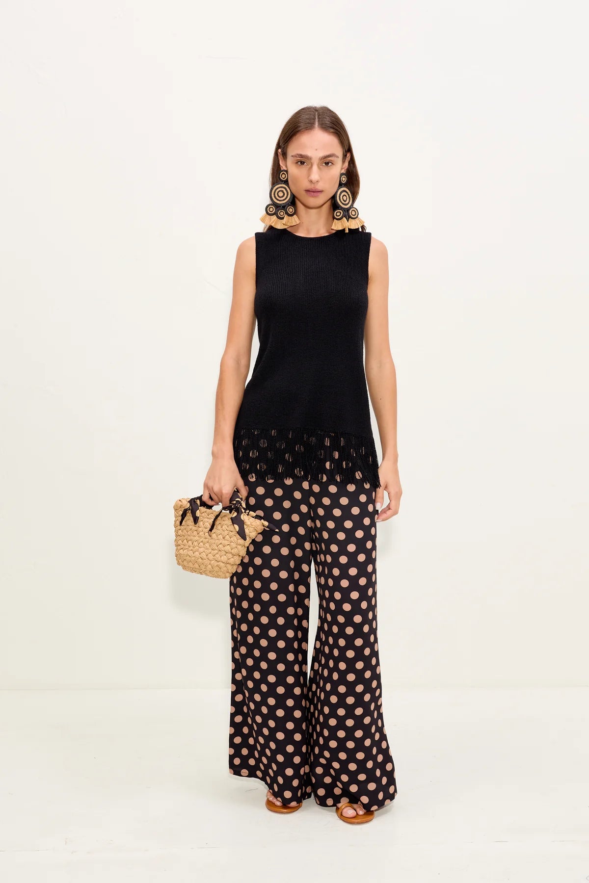Woman wearing a black sleeveless top and polka dot pants, holding a woven bag on a white background