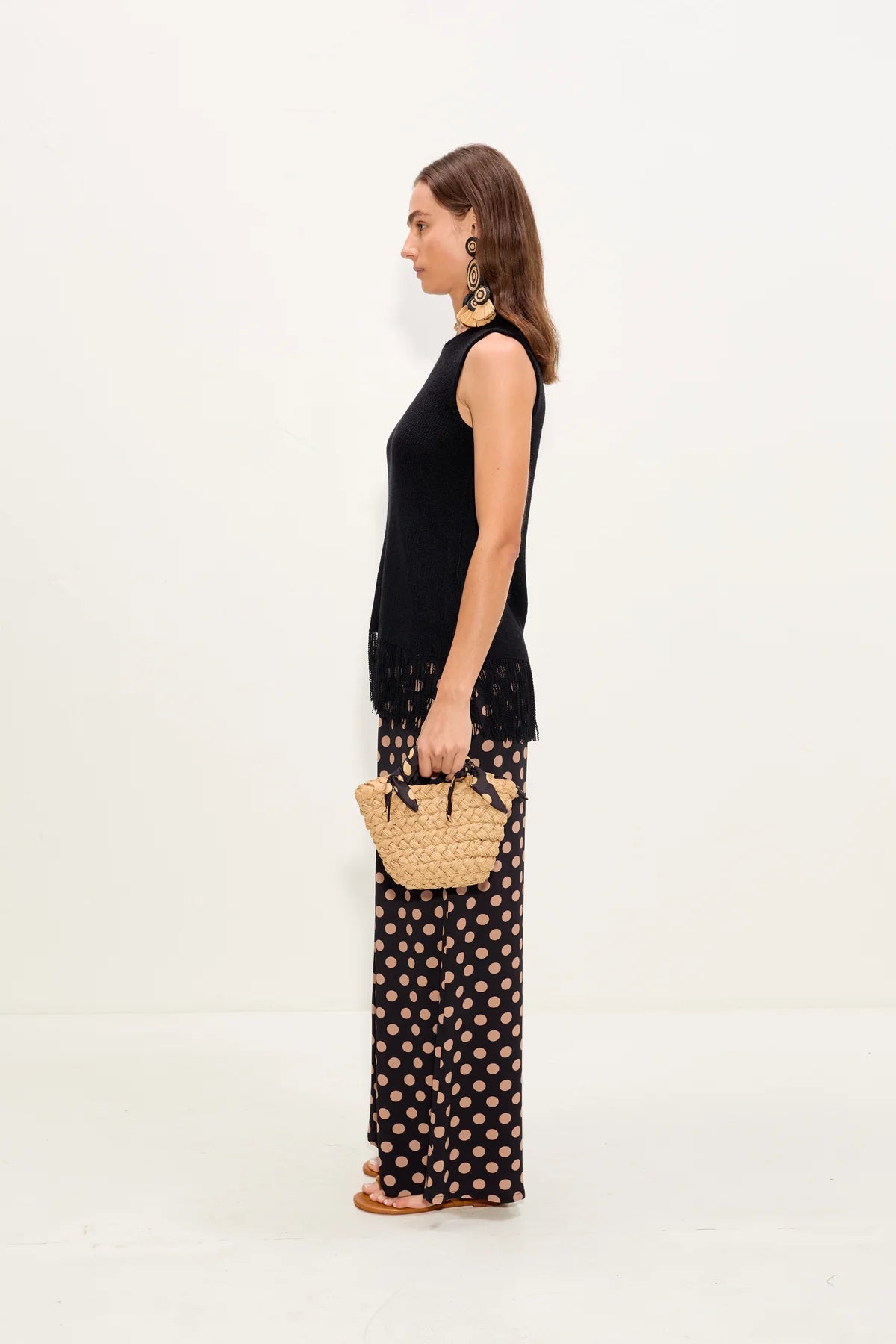 Woman holding a straw bag, wearing a black sleeveless top and patterned pants on a white background