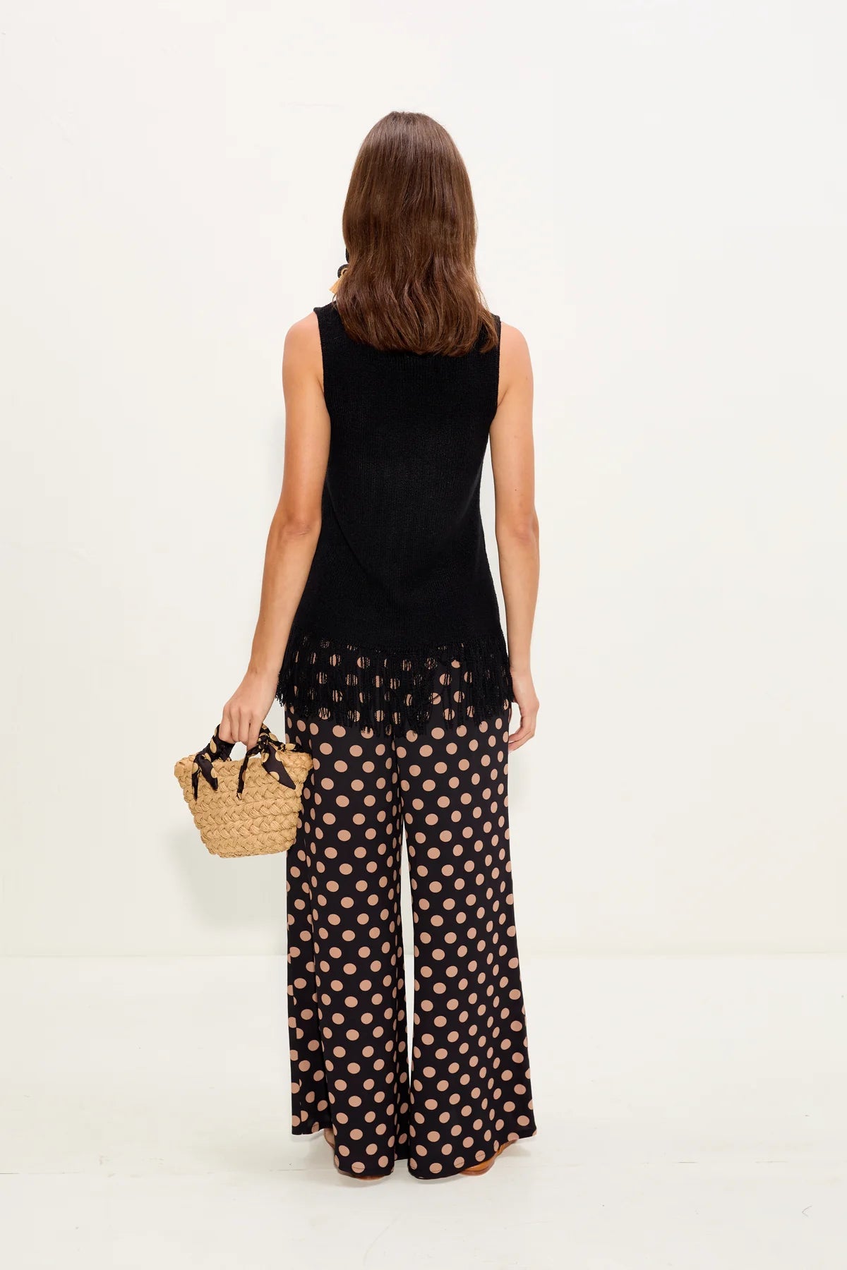 Woman wearing a black sleeveless top and patterned pants, holding a straw bag on a white background