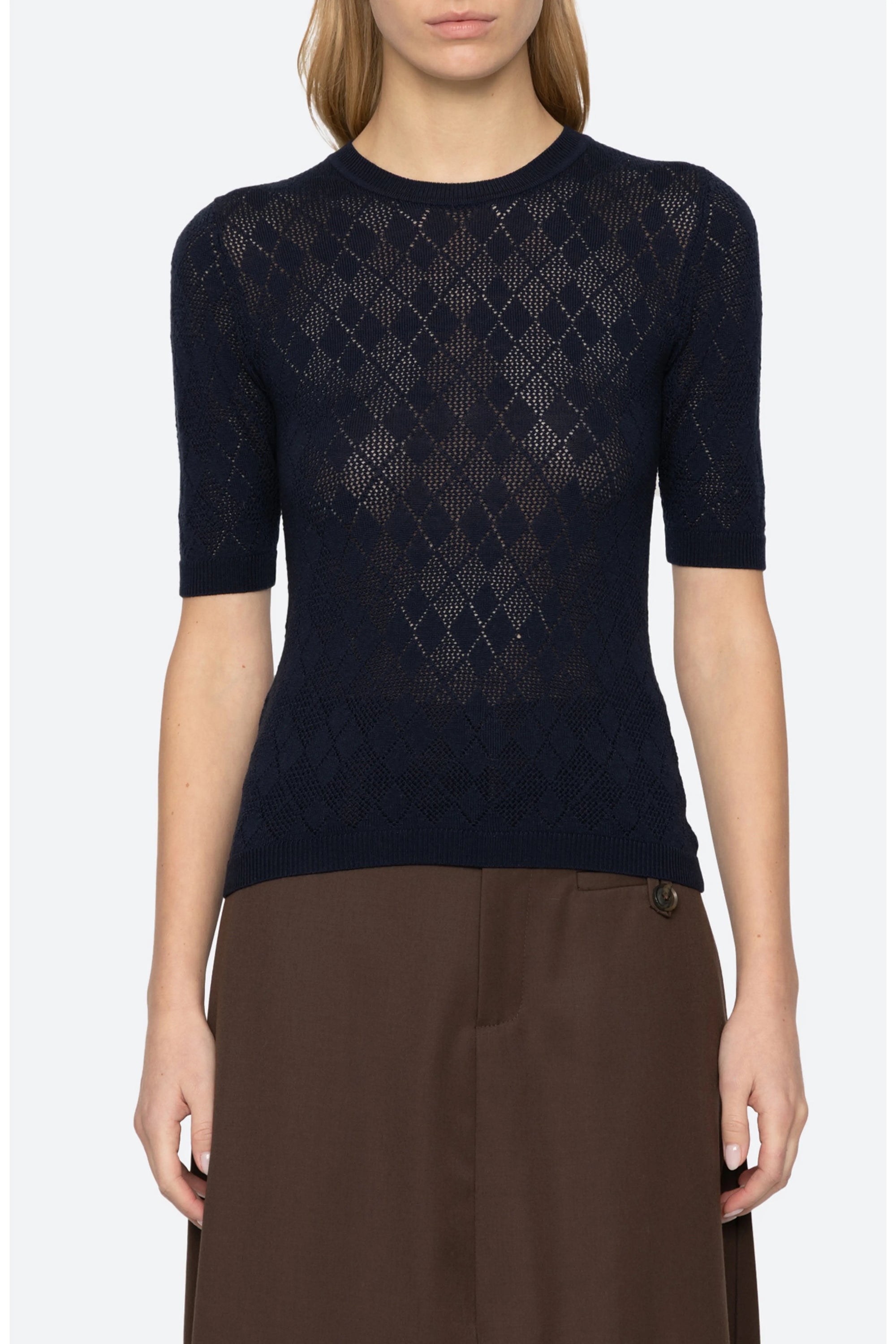 A woman wearing a navy blue short-sleeve knit top with an openwork argyle pattern, paired with brown trousers, against a light gray studio background.
