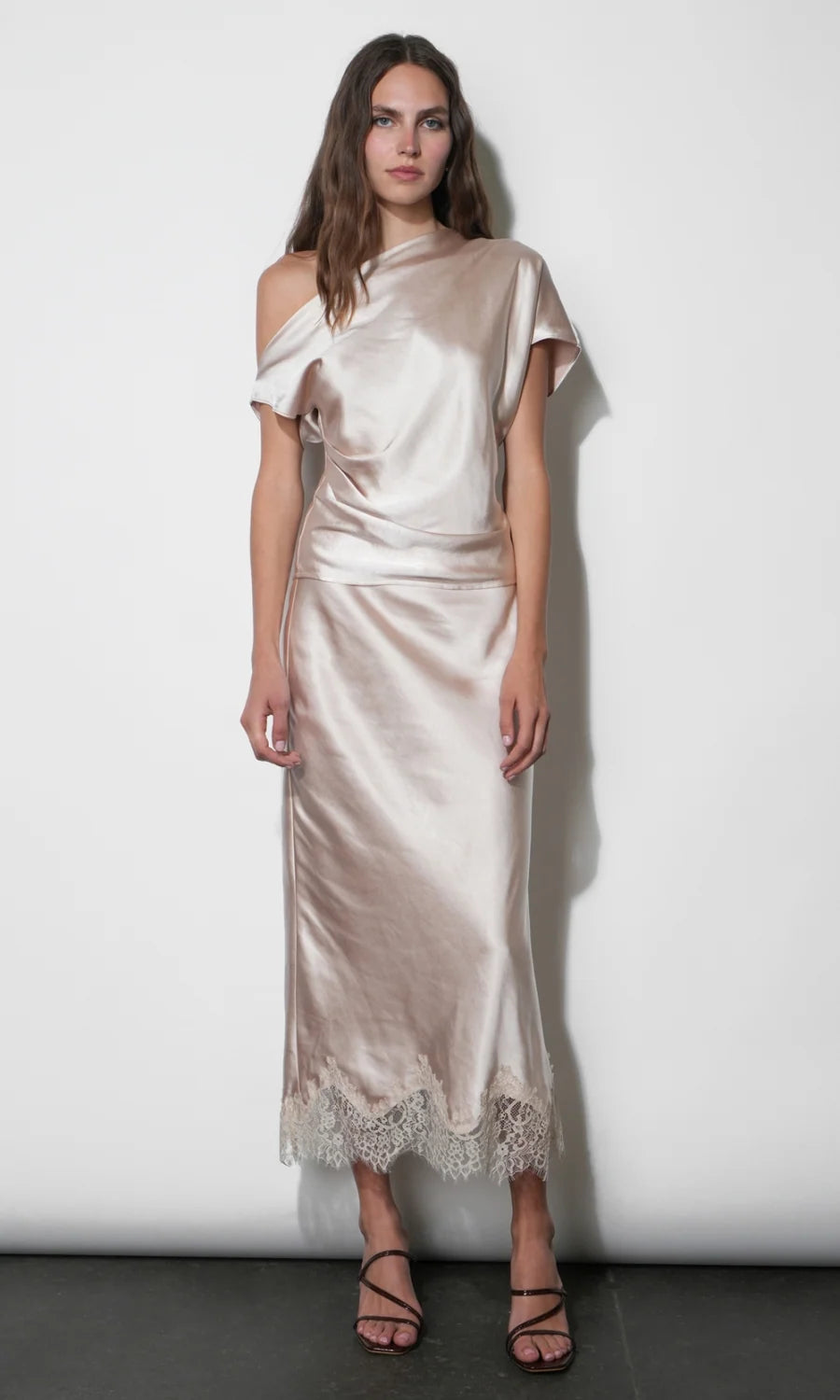 A woman models a champagne satin off-the-shoulder midi skirt with a lace hem, standing against a plain white studio wall.