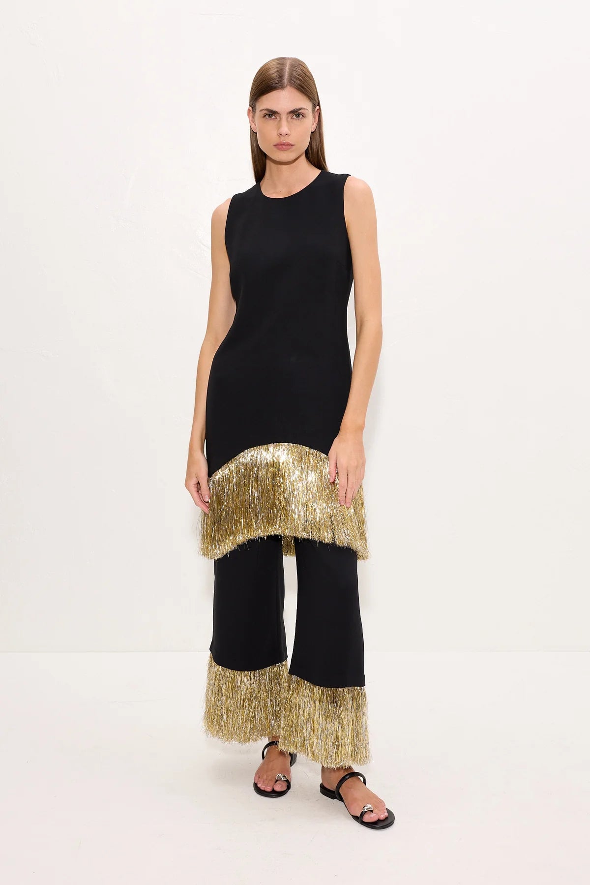 A model stands against a plain white studio background wearing a sleeveless black dress with a wide, textured metallic gold fringe hem and matching fringe along the bottom of wide-leg pants, styled with black sandals.