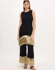 A model stands against a plain white studio background wearing a sleeveless black dress with a wide, textured metallic gold fringe hem and matching fringe along the bottom of wide-leg pants, styled with black sandals.