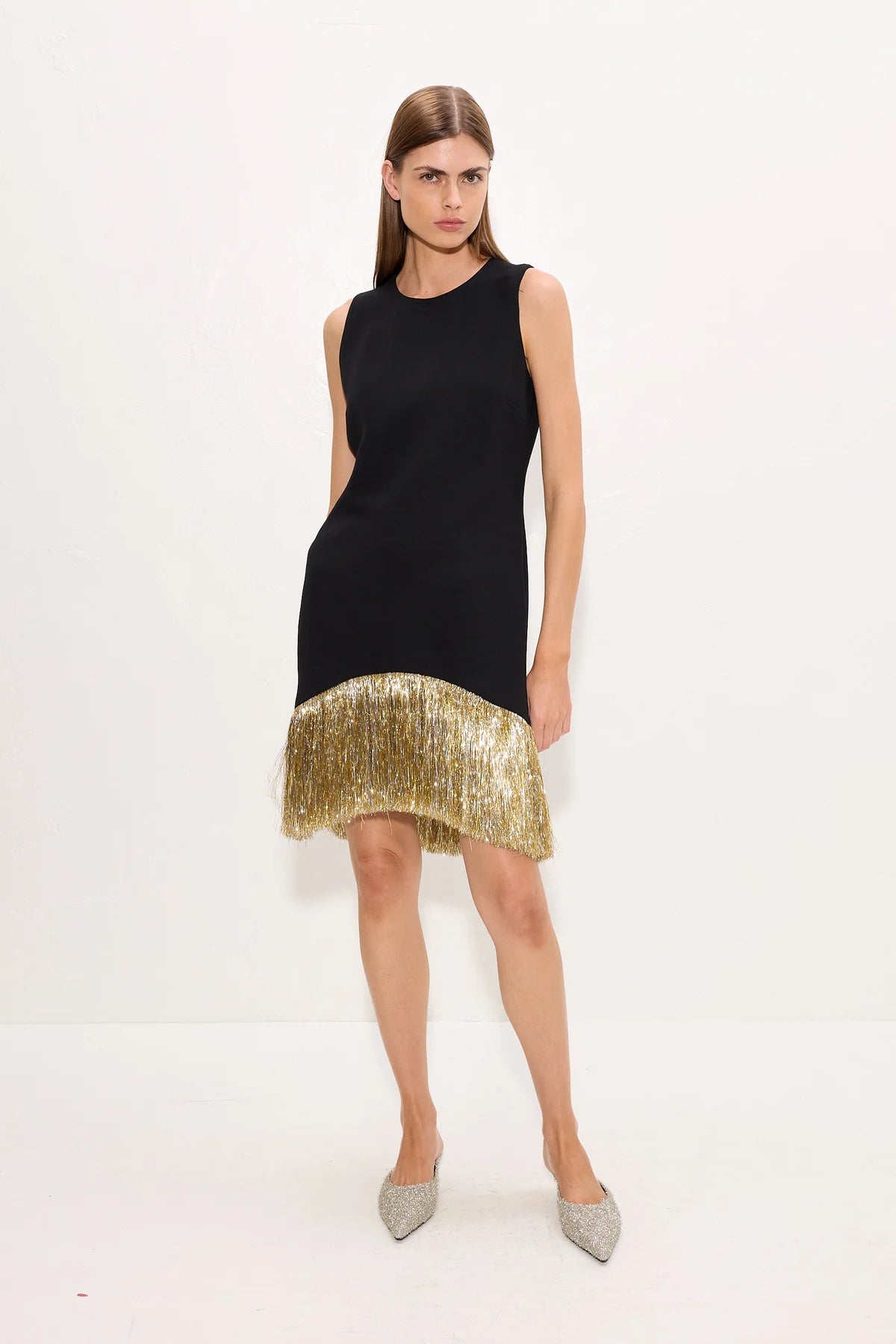 A woman models a sleeveless black mini dress with a gold hem, paired with silver pointed-toe heels, against a plain white studio background.