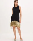 A woman models a sleeveless black mini dress with a gold hem, paired with silver pointed-toe heels, against a plain white studio background.