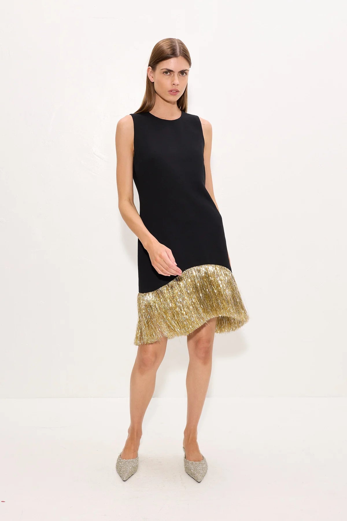 A woman modeling a sleeveless black dress with a flared gold hem, paired with pointed silver heels, against a plain white studio background.
