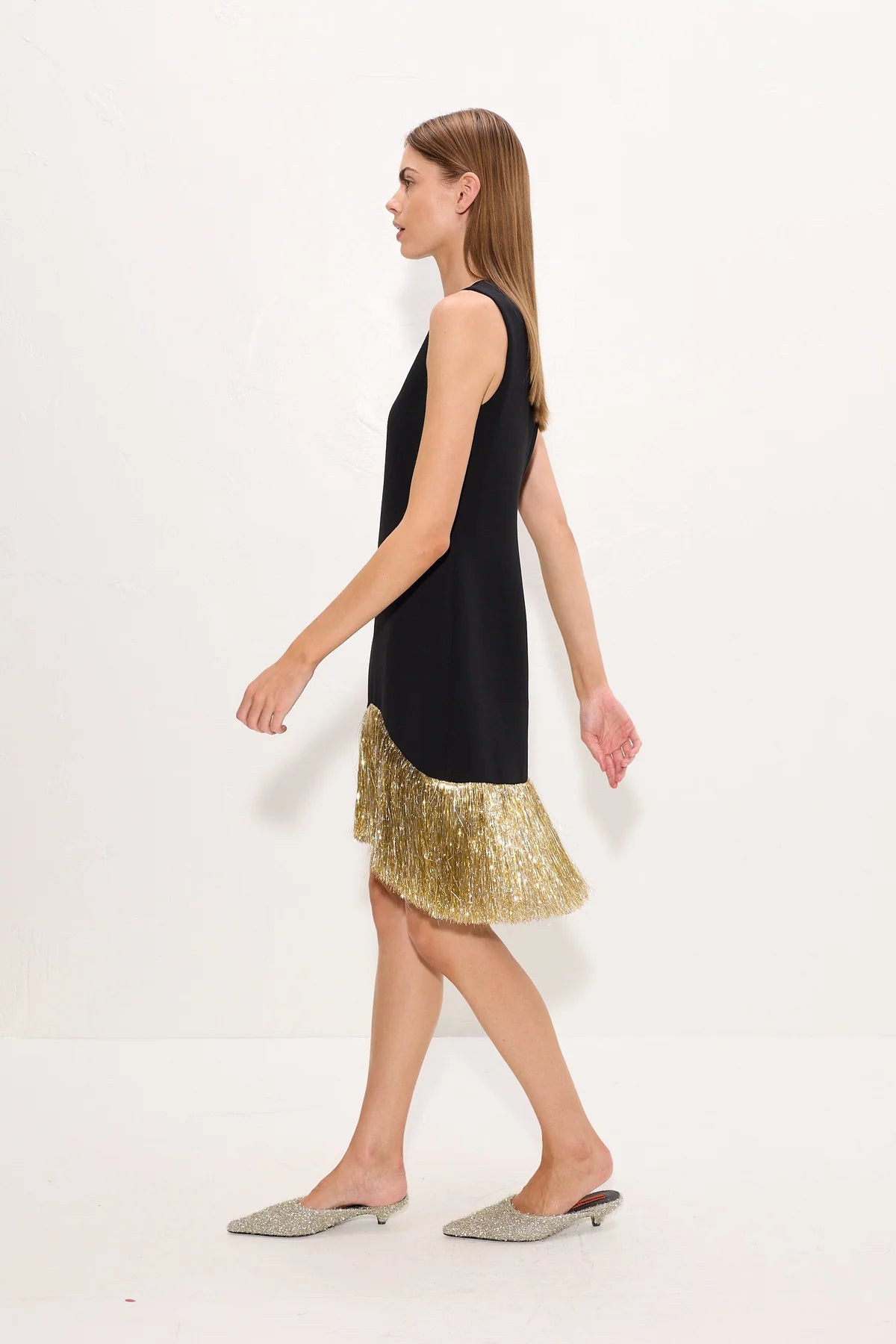 A woman models a sleeveless black dress with a flared hem covered in gold, paired with silver pointed flats, against a plain white studio background.