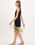 A woman models a sleeveless black dress with a flared hem covered in gold, paired with silver pointed flats, against a plain white studio background.