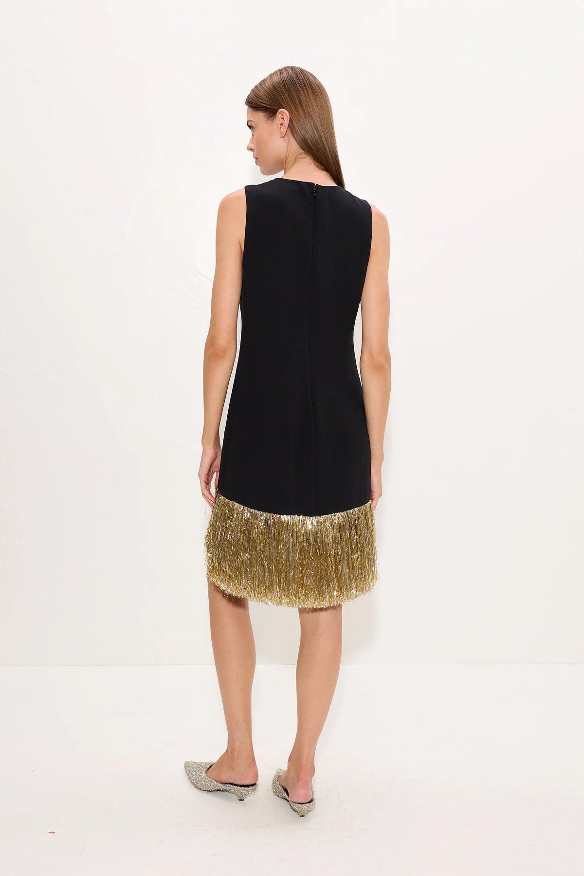A woman shown from the back wearing a sleeveless black dress with a textured, fringed gold hem, paired with gray pointed-toe mule heels, against a plain white studio background.