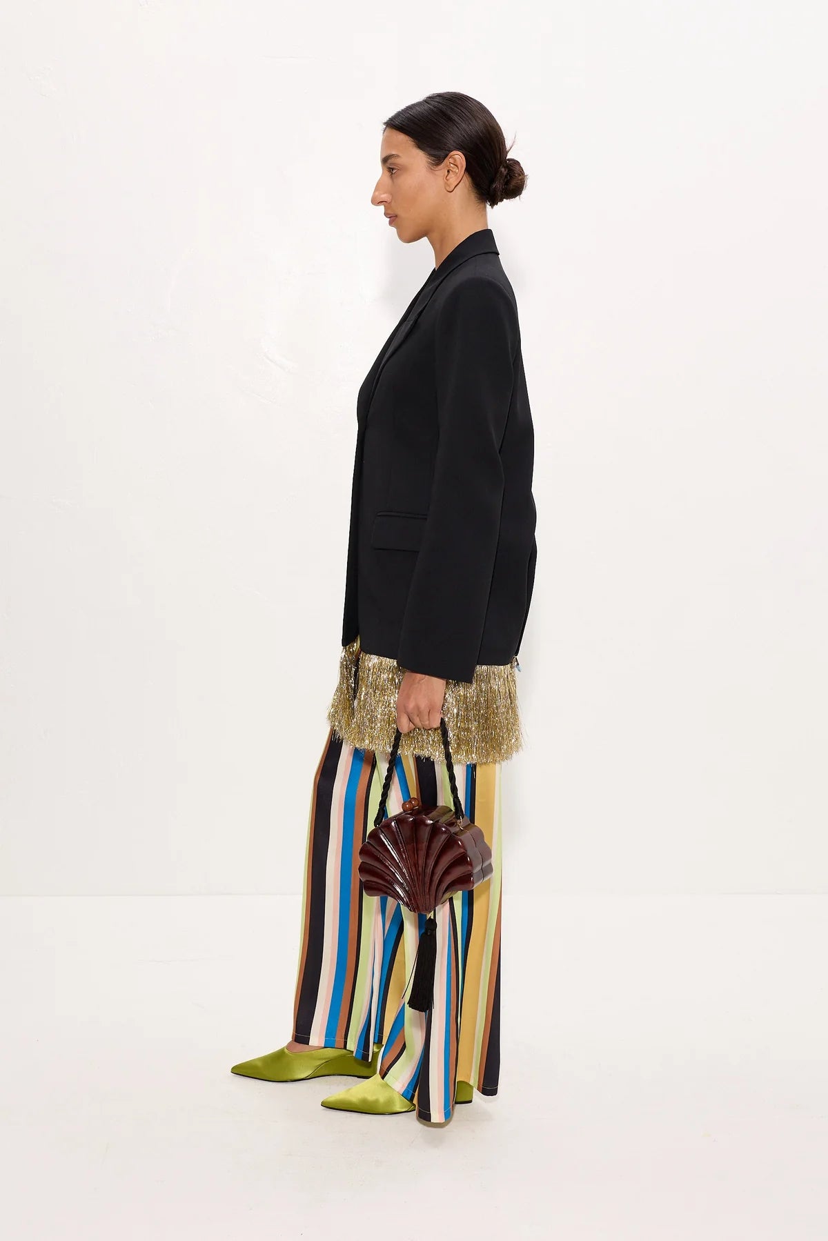 A model is shown in profile wearing a black blazer, wide-leg striped trousers in black, blue, yellow, and white, a textured gold skirt, and lime green pointed shoes, while holding a dark brown fan-shaped handbag against a plain white studio background.