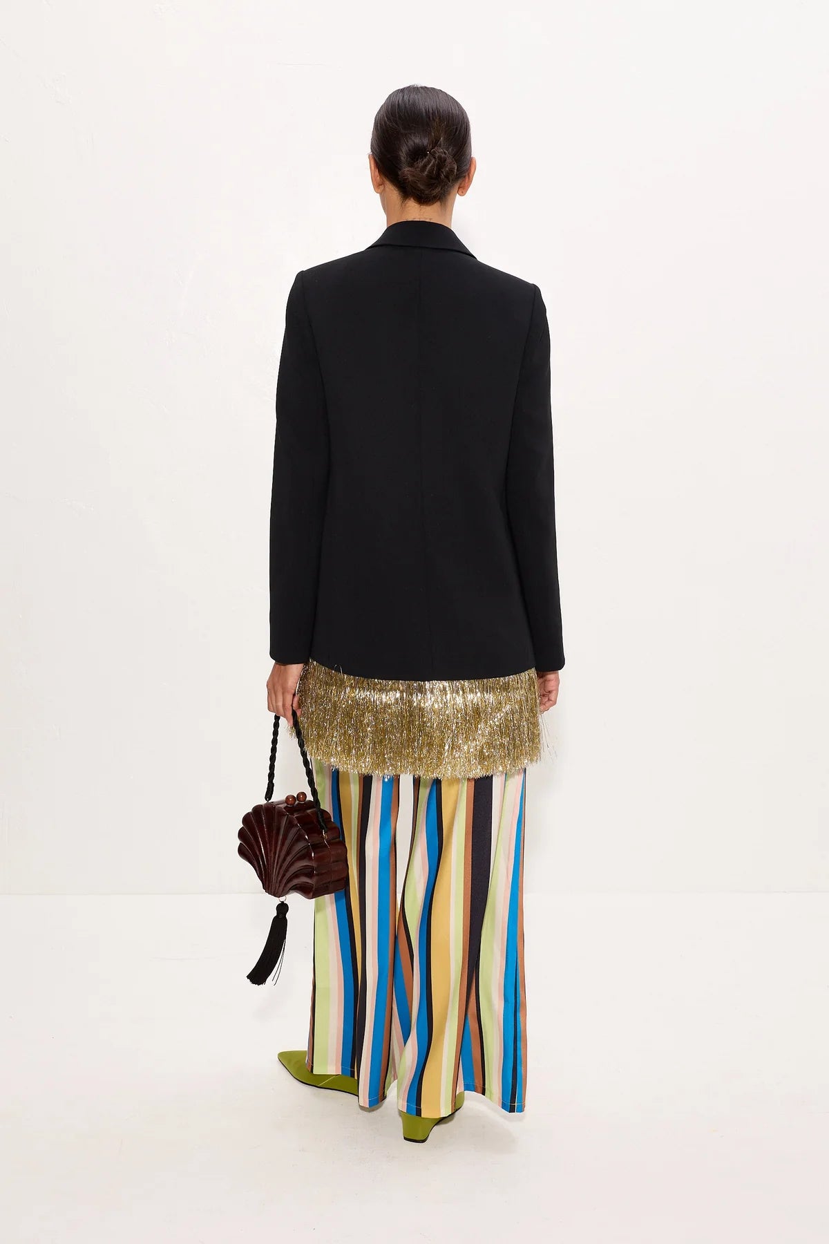 A model shown from the back wearing a black blazer, a gold sequined waistcoat, and a colorful vertically striped skirt in shades of blue, yellow, green, and brown. She is holding a dark brown handbag against a plain white studio background.
