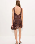 A woman viewed from behind wearing a sleeveless brown crochet knit dress with a low open back, holding a small brown handbag against a plain white studio background.