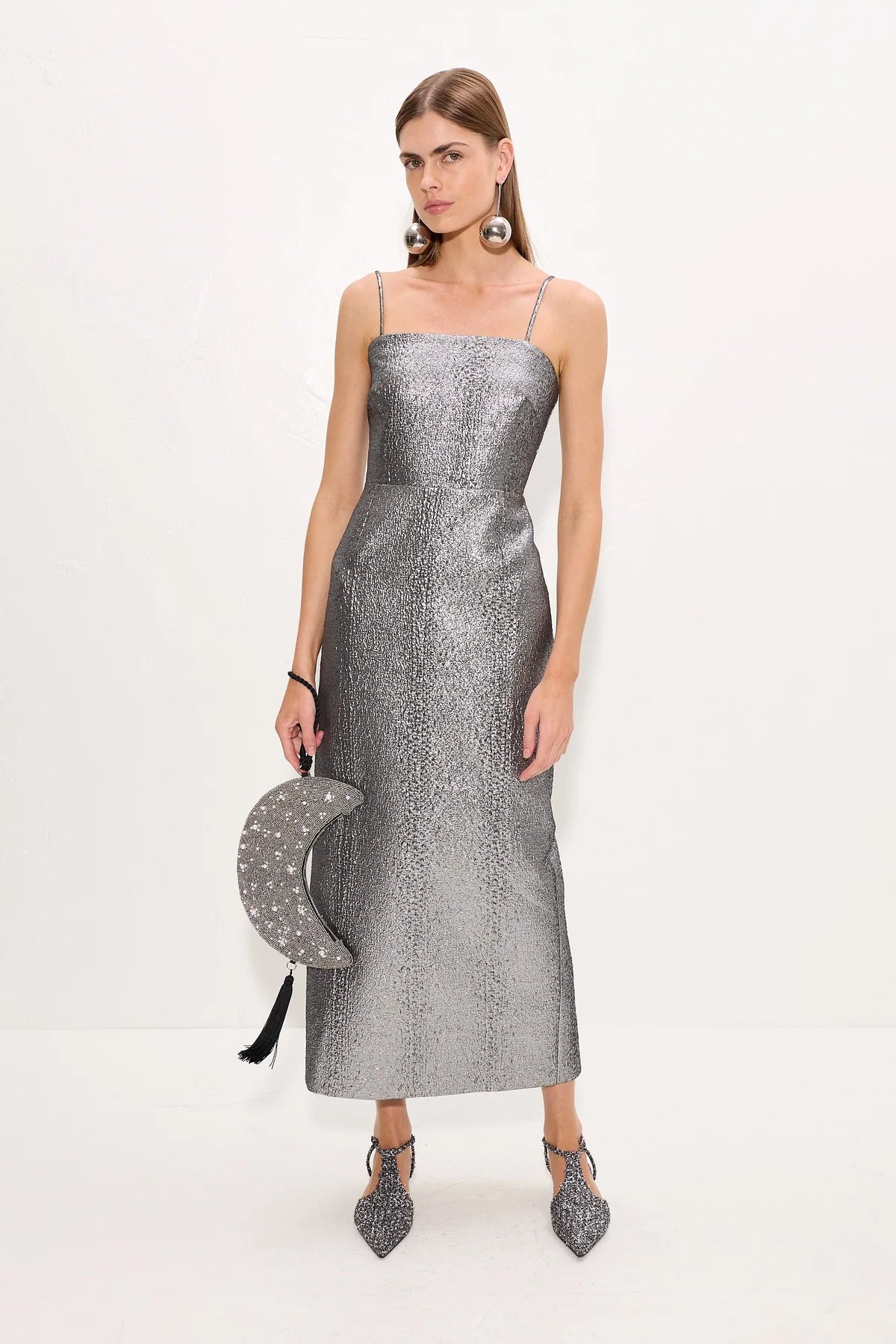 A model wearing a metallic silver spaghetti-strap midi dress, holding a matching silver crescent moon clutch bag with black tassels, and wearing silver pointed-toe heels against a plain white studio background.