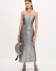 A model wearing a metallic silver spaghetti-strap midi dress, holding a matching silver crescent moon clutch bag with black tassels, and wearing silver pointed-toe heels against a plain white studio background.
