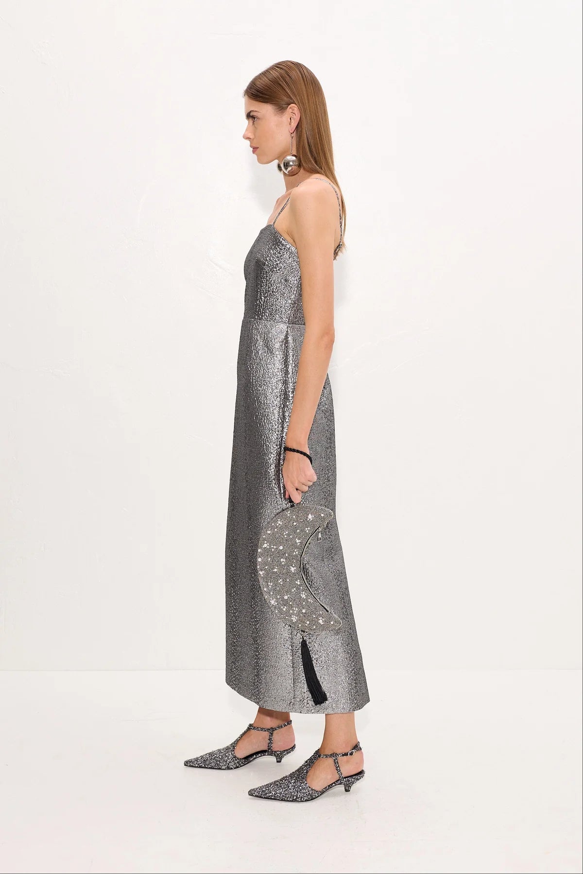 A woman shown from the side wearing a metallic silver spaghetti-strap midi dress with a textured pattern, holding a matching silver clutch, against a plain white studio background.