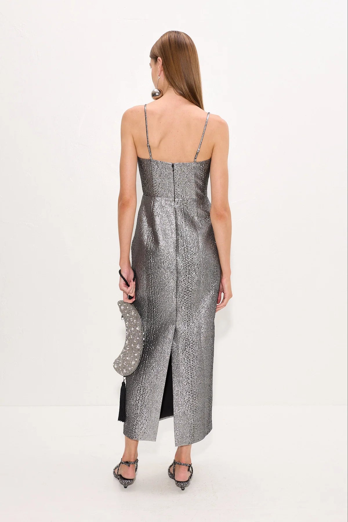 A woman shown from the back wearing a metallic silver spaghetti-strap midi dress with a textured pattern, holding a matching silver clutch, against a plain white studio background.