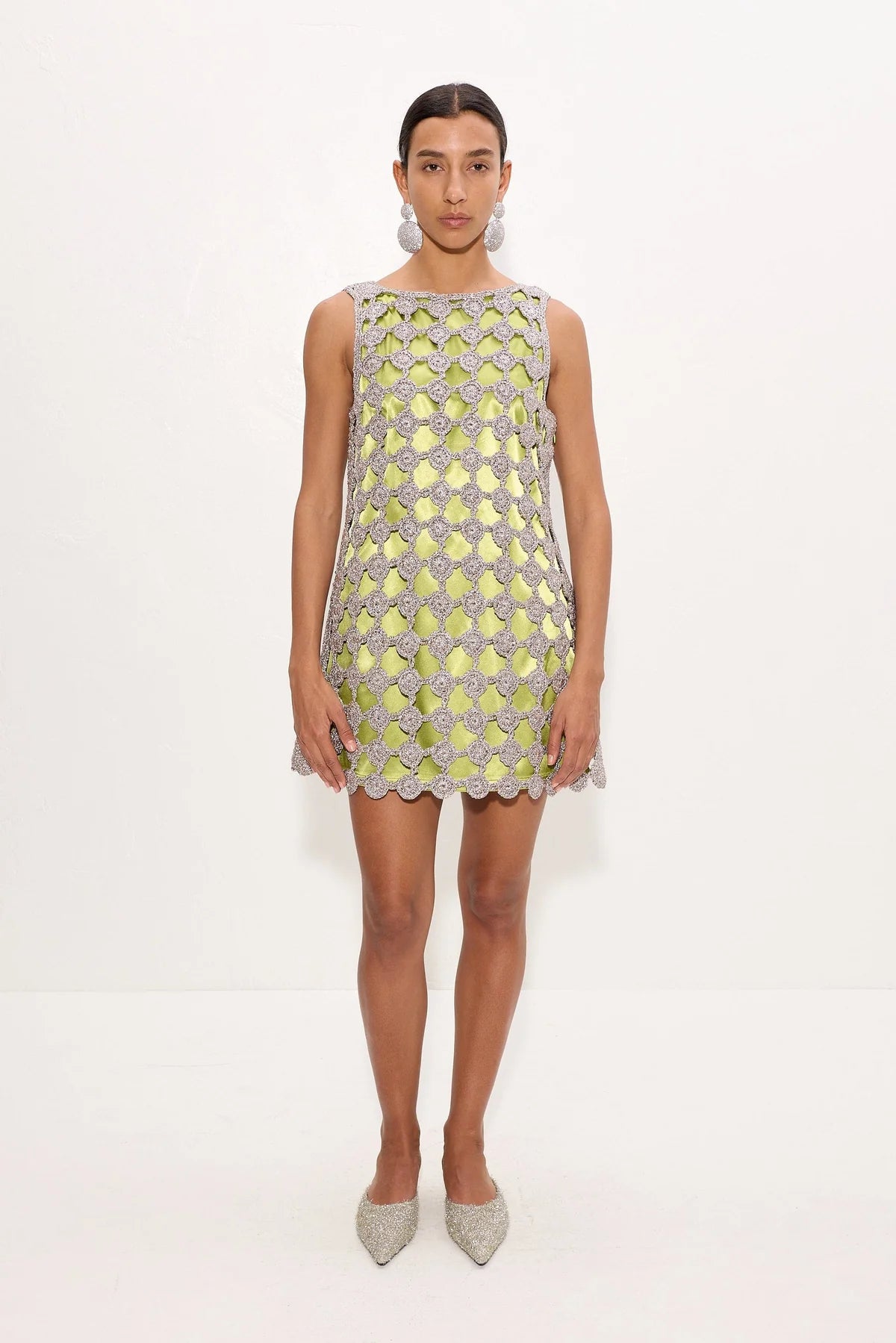 A model stands against a plain white studio background wearing a sleeveless, short dress covered in a metallic silver and pale yellow-green geometric mesh pattern with scalloped edges, paired with sparkly silver pointed shoes.