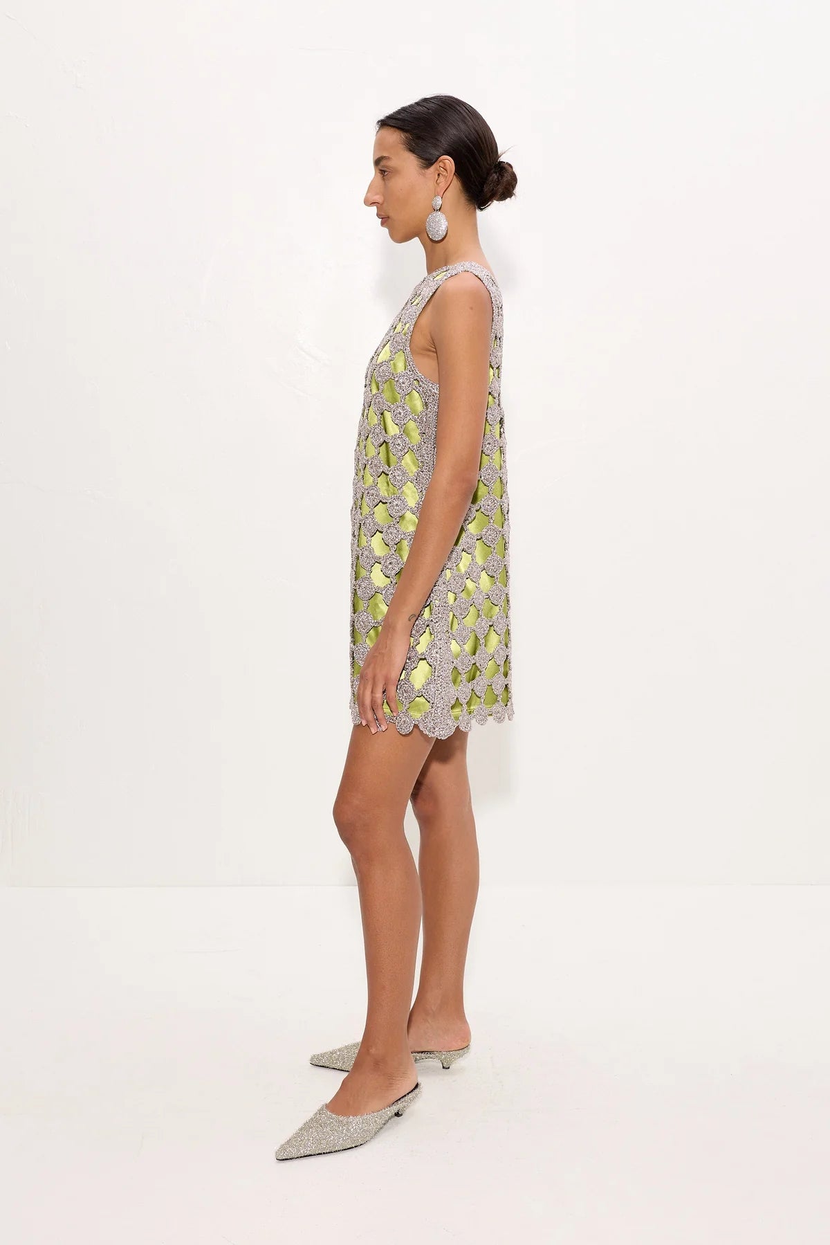 A woman stands in profile wearing a sleeveless, short silver dress with a green and yellow floral pattern and scalloped edges, paired with silver pointed flats, against a plain white studio background.
