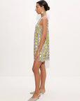 A woman stands in profile wearing a sleeveless, short silver dress with a green and yellow floral pattern and scalloped edges, paired with silver pointed flats, against a plain white studio background.