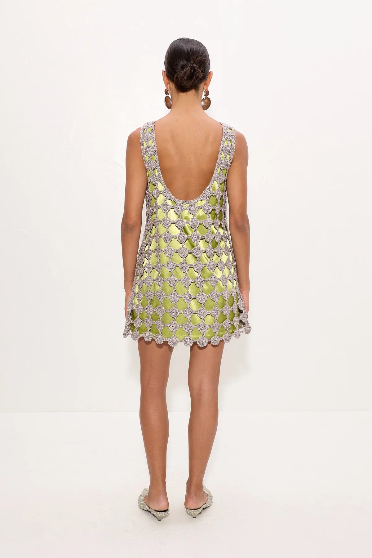 A woman viewed from the back wearing a sleeveless open-back mini dress covered in metallic gold and silver circular embellishments, set against a plain white studio background.