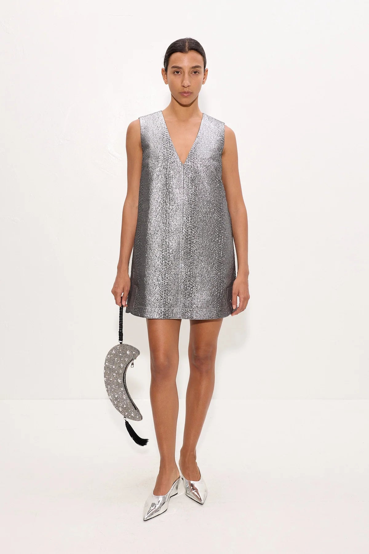 A model stands against a plain white studio background wearing a sleeveless silver mini dress with a deep V-neckline and holding a matching silver sequined crescent-shaped handbag.