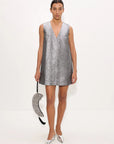 A model stands against a plain white studio background wearing a sleeveless silver mini dress with a deep V-neckline and holding a matching silver sequined crescent-shaped handbag.