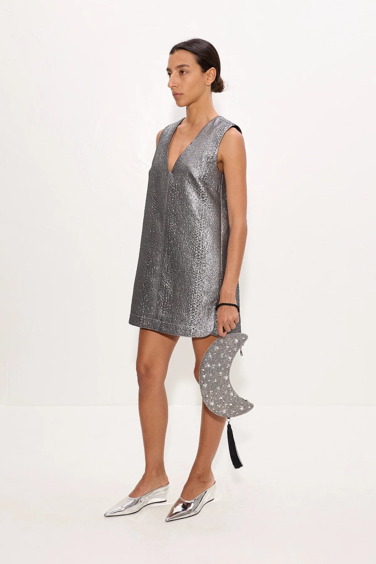 A woman models a sleeveless silver mini dress with a deep V-neck, paired with silver pointed-toe flats and a matching silver star-patterned crescent moon clutch bag, set against a plain white studio background.