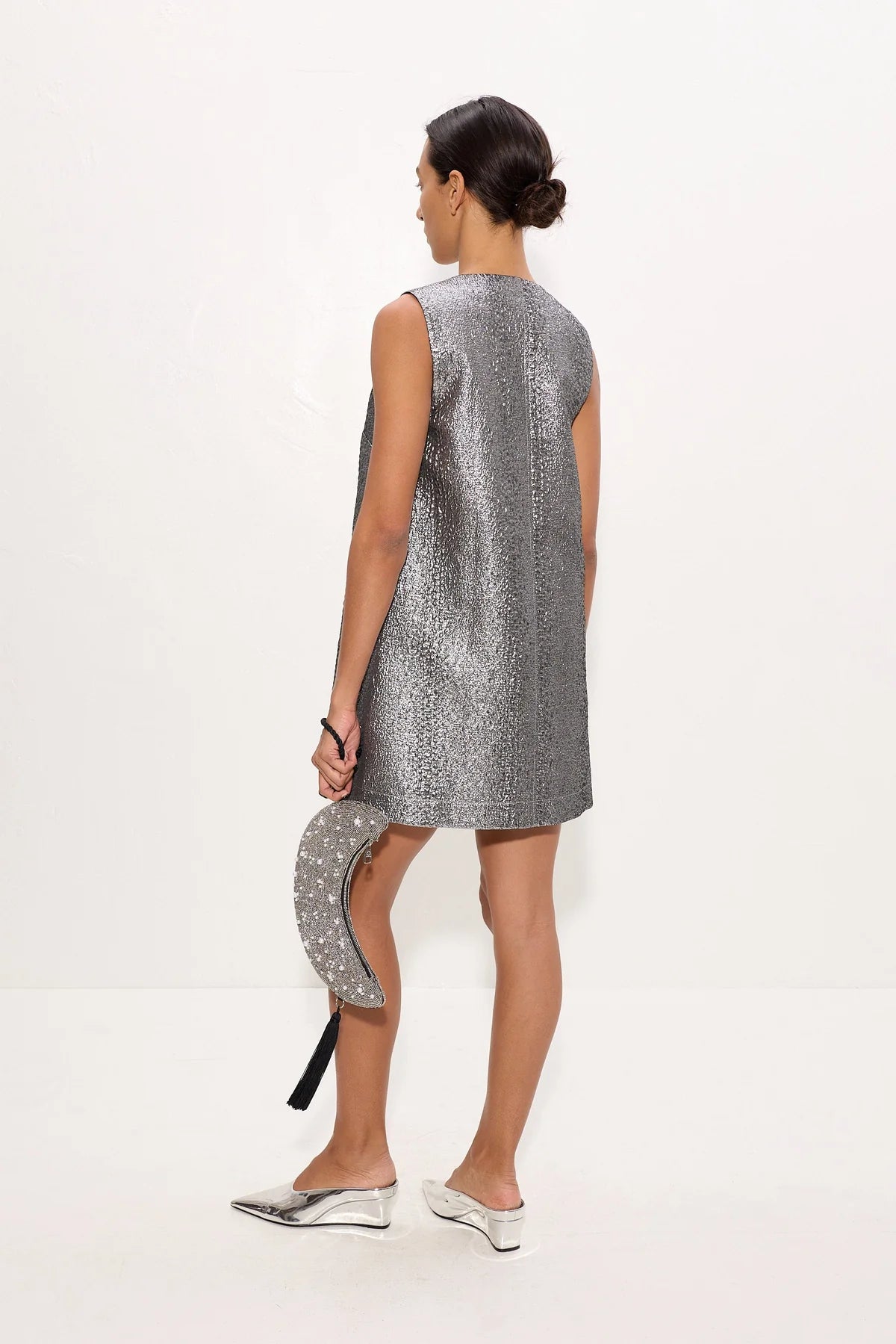 A woman shown from the back wearing a sleeveless silver textured mini dress, holding a matching silver patterned clutch, and wearing silver pointed flats against a plain white studio background.