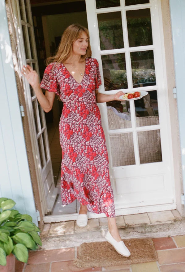 A woman wearing a red and white floral midi dress with short flutter sleeves and a V-neckline, standing in a doorway and holding a plate of tomatoes.