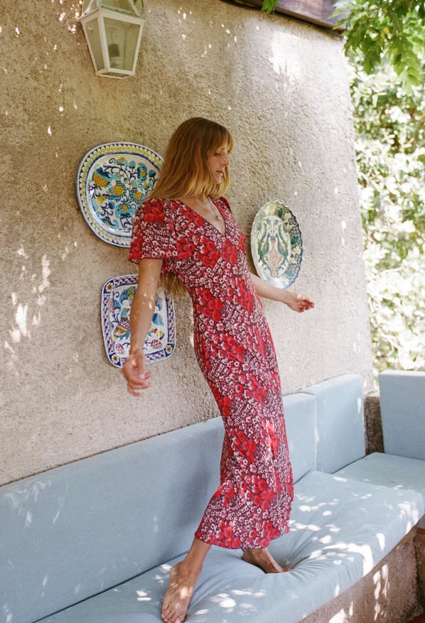 A woman wearing a red and white floral midi dress stands barefoot on a light blue outdoor bench against a textured beige stucco wall with decorative plates.