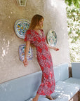 A woman wearing a red and white floral midi dress stands barefoot on a light blue outdoor bench against a textured beige stucco wall with decorative plates.