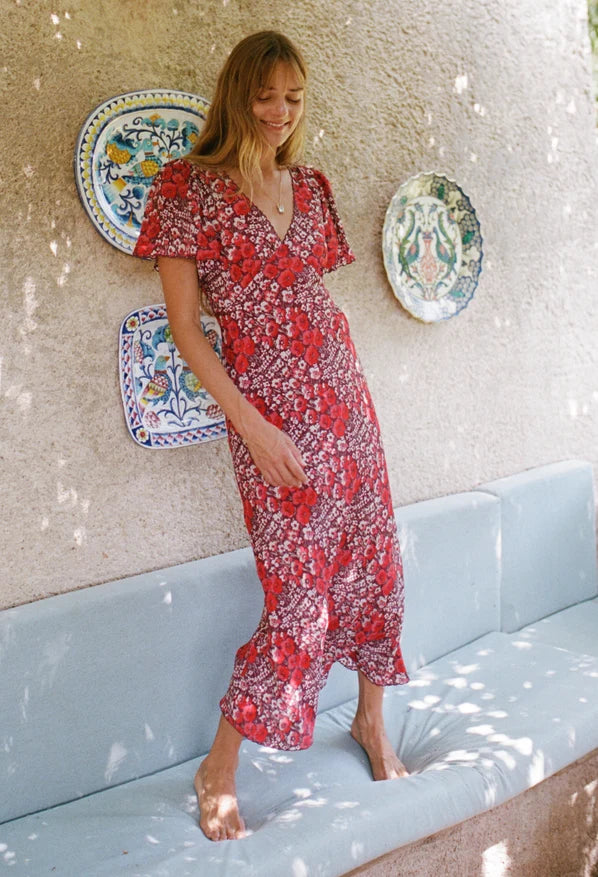 A woman stands barefoot on a light blue cushioned bench against a textured beige wall, wearing a red and white floral print midi dress with flutter sleeves and a V-neckline. Decorative ceramic plates hang on the wall behind her.