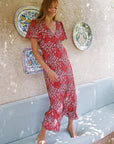 A woman stands barefoot on a light blue cushioned bench against a textured beige wall, wearing a red and white floral print midi dress with flutter sleeves and a V-neckline. Decorative ceramic plates hang on the wall behind her.