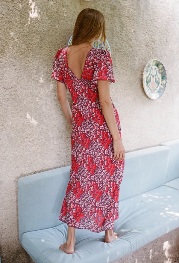 A woman stands barefoot on a light blue cushioned bench, viewed from behind, wearing a red and white floral maxi dress with a deep open back and short flutter sleeves. The background features a textured beige stucco wall with a decorative plate.