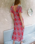 A woman stands barefoot on a light blue cushioned bench, viewed from behind, wearing a red and white floral maxi dress with a deep open back and short flutter sleeves. The background features a textured beige stucco wall with a decorative plate.