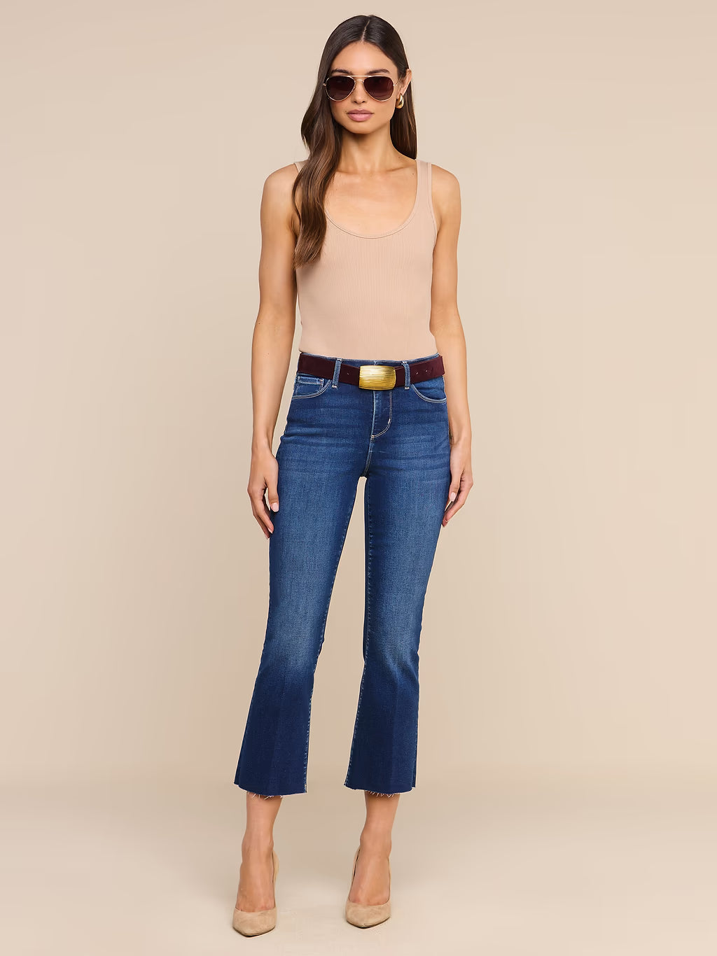 A woman modeling a pair of dark blue cropped flare jeans with a brown belt and gold buckle, paired with a beige tank top and nude heels, against a solid beige background.