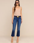 A woman modeling a pair of dark blue cropped flare jeans with a brown belt and gold buckle, paired with a beige tank top and nude heels, against a solid beige background.