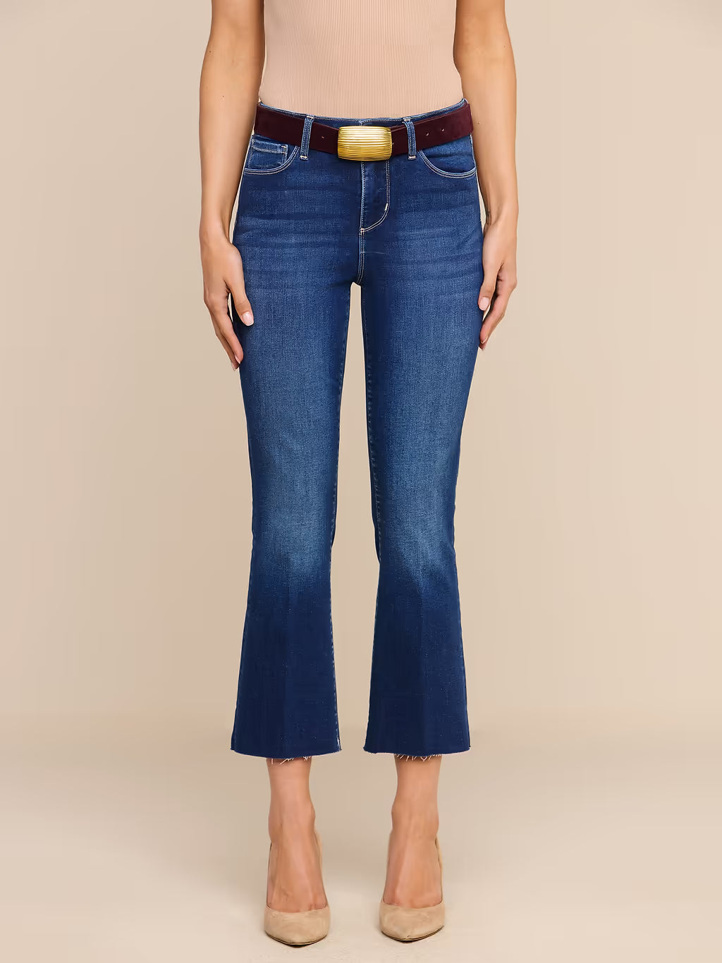 A model wearing blue cropped flare jeans with a brown belt and gold buckle, paired with beige heels, against a solid beige background.