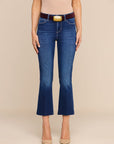 A model wearing blue cropped flare jeans with a brown belt and gold buckle, paired with beige heels, against a solid beige background.