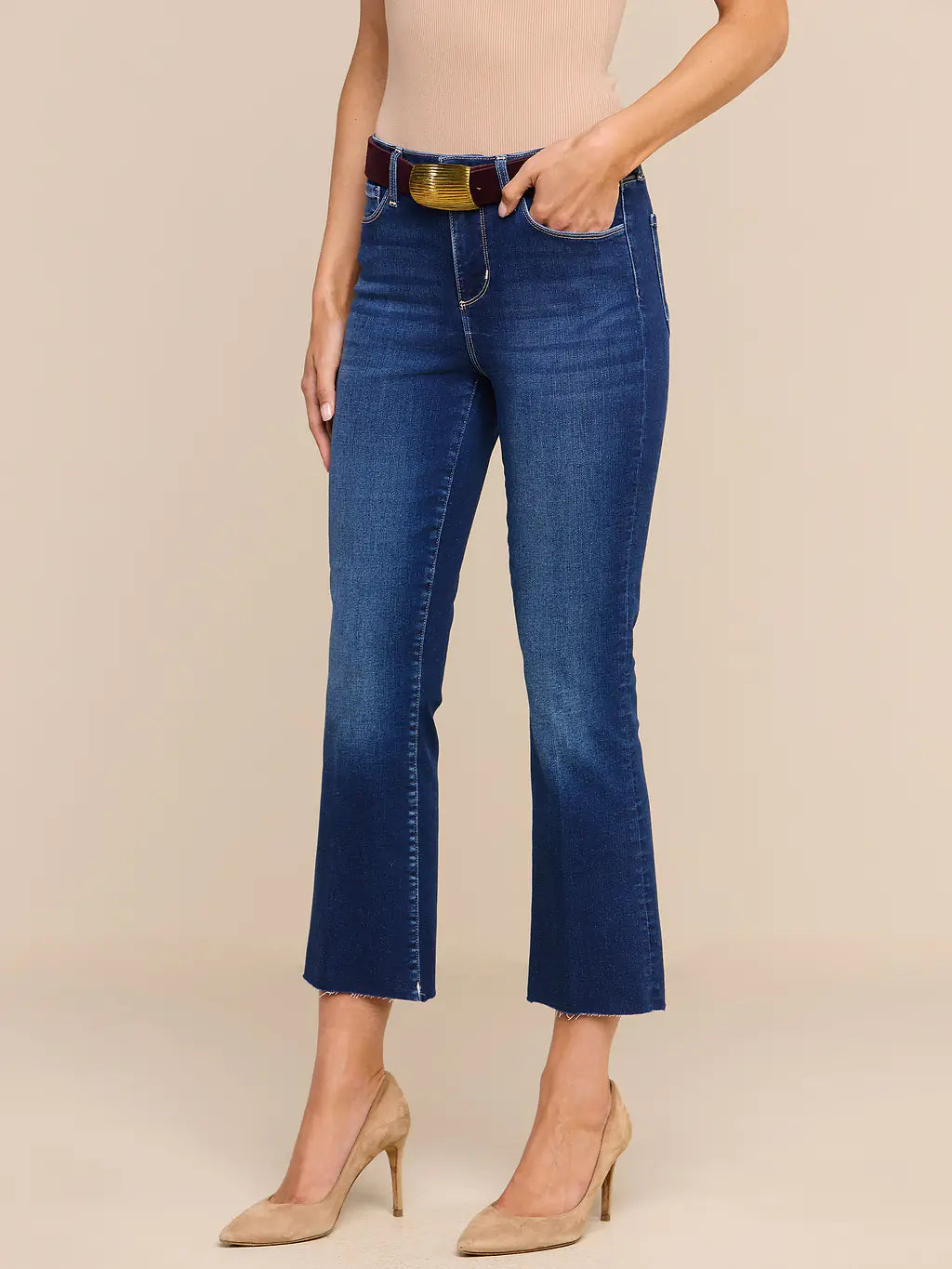 A model wearing dark blue cropped flare jeans with a gold-tone belt, paired with beige heels and a beige top, standing against a solid beige studio background.