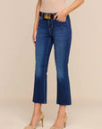 A model wearing dark blue cropped flare jeans with a gold-tone belt, paired with beige heels and a beige top, standing against a solid beige studio background.