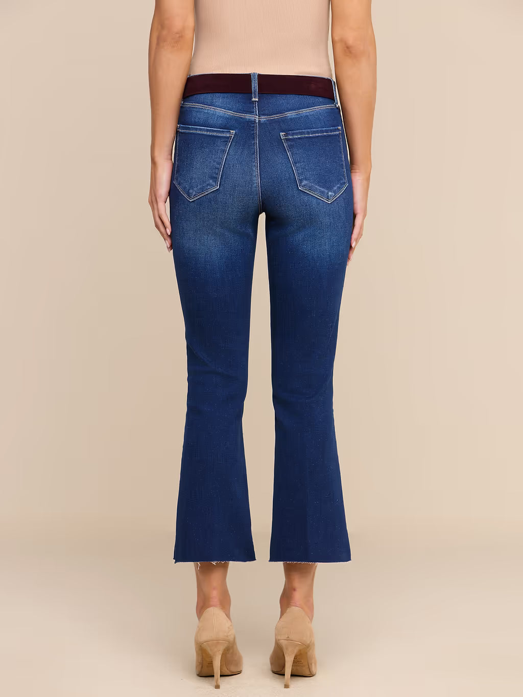 Back view of a model wearing blue flare jeans with a dark brown belt, paired with beige high heels, against a solid beige background.