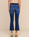 Back view of a model wearing blue flare jeans with a dark brown belt, paired with beige high heels, against a solid beige background.